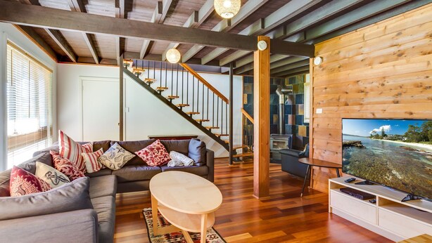 A cozy living room with a wooden staircase, a sofa with patterned cushions, a TV, and a view of the sea through a window. - Shanti at Hyams Beach, Jervis Bay (Hyams Beach)