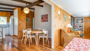 A cozy room with a wooden interior, a dining table with chairs, a bookshelf, and a bed with a floral bedspread. - Shanti at Hyams Beach, Jervis Bay (Hyams Beach)