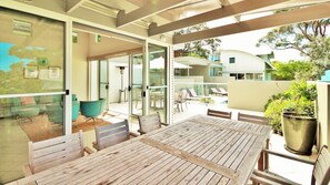 Ein Holztisch mit Stühlen in einem überdachten Außenbereich, mit Glastüren zu einem Pool und Liegestühlen.