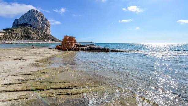 Uma praia com águas cristalinas, um afloramento rochoso e uma grande formação rochosa ao fundo.