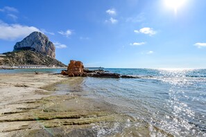 A beach with clear water, a rocky outcrop, and a large rock formation in the background.