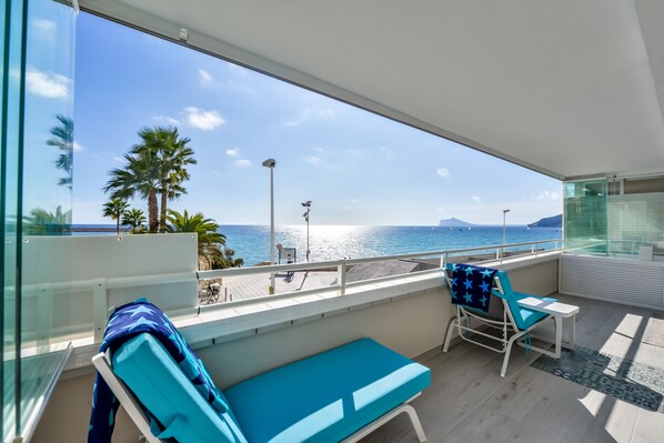 A balcony with a view of the sea, a lounge chair, a small table, and a bench.