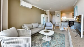 Uma sala de estar moderna com sofá, poltronas, uma mesa de centro e uma área de cozinha com mesa e cadeiras.
