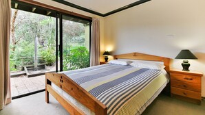 Un dormitorio con una cama grande, cabecera de madera, una mesita de noche con lámpara y vistas a zonas verdes a través de una puerta corrediza de vidrio.