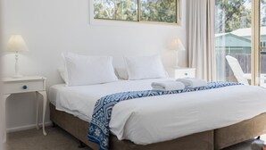 A neatly made bed with white linens and a blue patterned throw, two bedside tables with lamps, and a window with curtains.