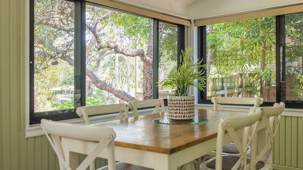 A dining area with a wooden table and white chairs, large windows with a view of trees and a fence, and a potted plant on the table.