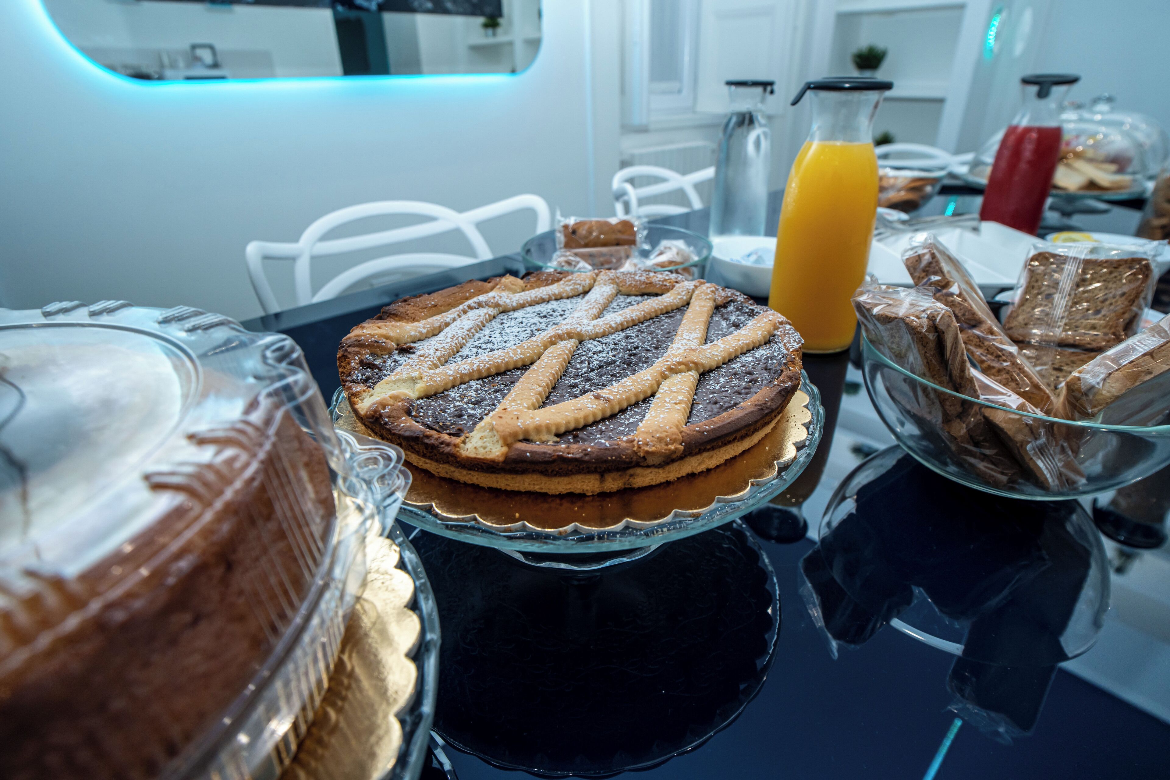 Un bufé de desayuno con diversos pasteles, una tarta y un vaso de zumo de naranja sobre una mesa negra reflectante.