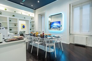 Una cocina moderna con armarios blancos, una zona de comedor con sillas blancas y un televisor de pantalla plana grande montado en la pared.