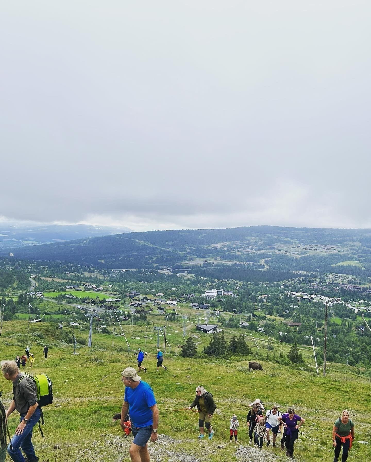 Un paysage vallonné avec une petite ville qui se dessine à l’horizon, un ciel dégagé et des gens qui se promènent.