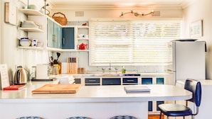 A kitchen with a central island, built-in shelves, and a window with blinds. - The Shack at Palm Beach (Sanctuary Point)
