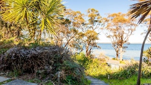 A coastal landscape with a variety of trees and shrubs, a clear view of the sea, and a paved path leading towards the water. - The Shack at Palm Beach (Sanctuary Point)