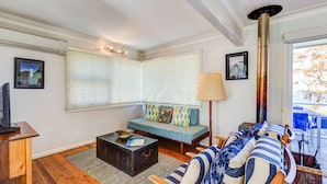 A cozy living room with a sofa, a coffee table, and a lamp. There is a window with blinds, a fireplace, and a view of the outdoors. - The Shack at Palm Beach (Sanctuary Point)
