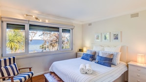 A bedroom with a large window, a bed with blue and white bedding, a chair, and a bedside table with a lamp. - The Shack at Palm Beach (Sanctuary Point)