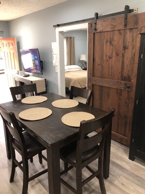 Un comedor con una mesa de madera oscura y cuatro sillas, cada una con un mantel tejido. Se observa una puerta corrediza de estilo rústico al fondo.