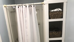 A shower area with a white shower curtain, a white built-in shelf with baskets, and a wooden floor. - The Cottage at Sunset View Estate.  Close to rafting, hiking, Dollywood 30 mins. (Cosby)