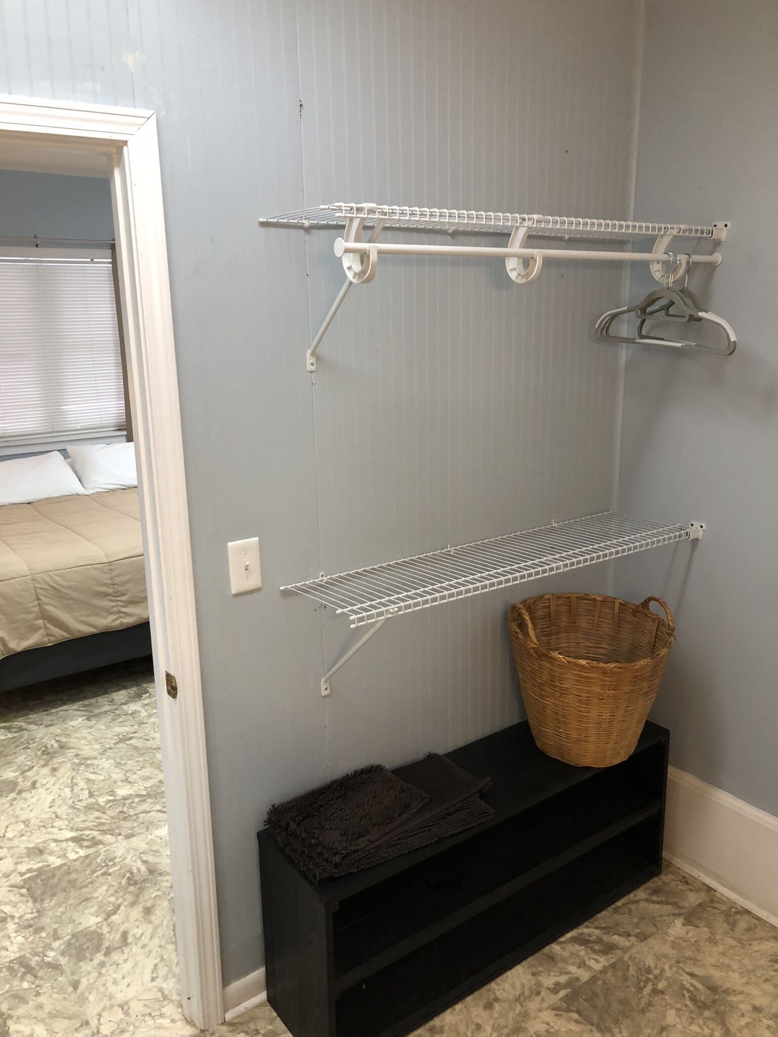 A room with a grey wall, a white door, a bed with a beige bedspread, and two white wire shelves with a wicker basket on a black shelf.