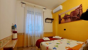 Ein Hotelzimmer mit Bett, Fenster mit durchsichtigen Vorhängen, Klimaanlage, Wandregal und einem Stadtbild.