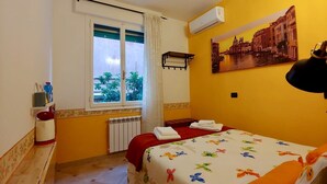 Ein Hotelzimmer mit Bett, einem fenster mit Vorhängen, einem Heizkörper, einem Schreibtisch mit Stuhl, einer Wand-Klimaanlage und einem Stadtbild.