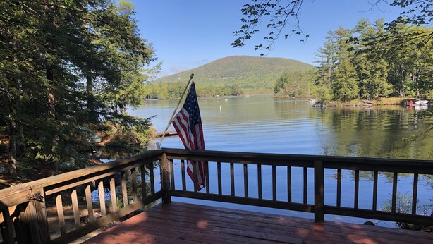 Un pontile di legno con una bandiera americana, affacciato su un lago tranquillo con barche e una collina boscosa sullo sfondo.