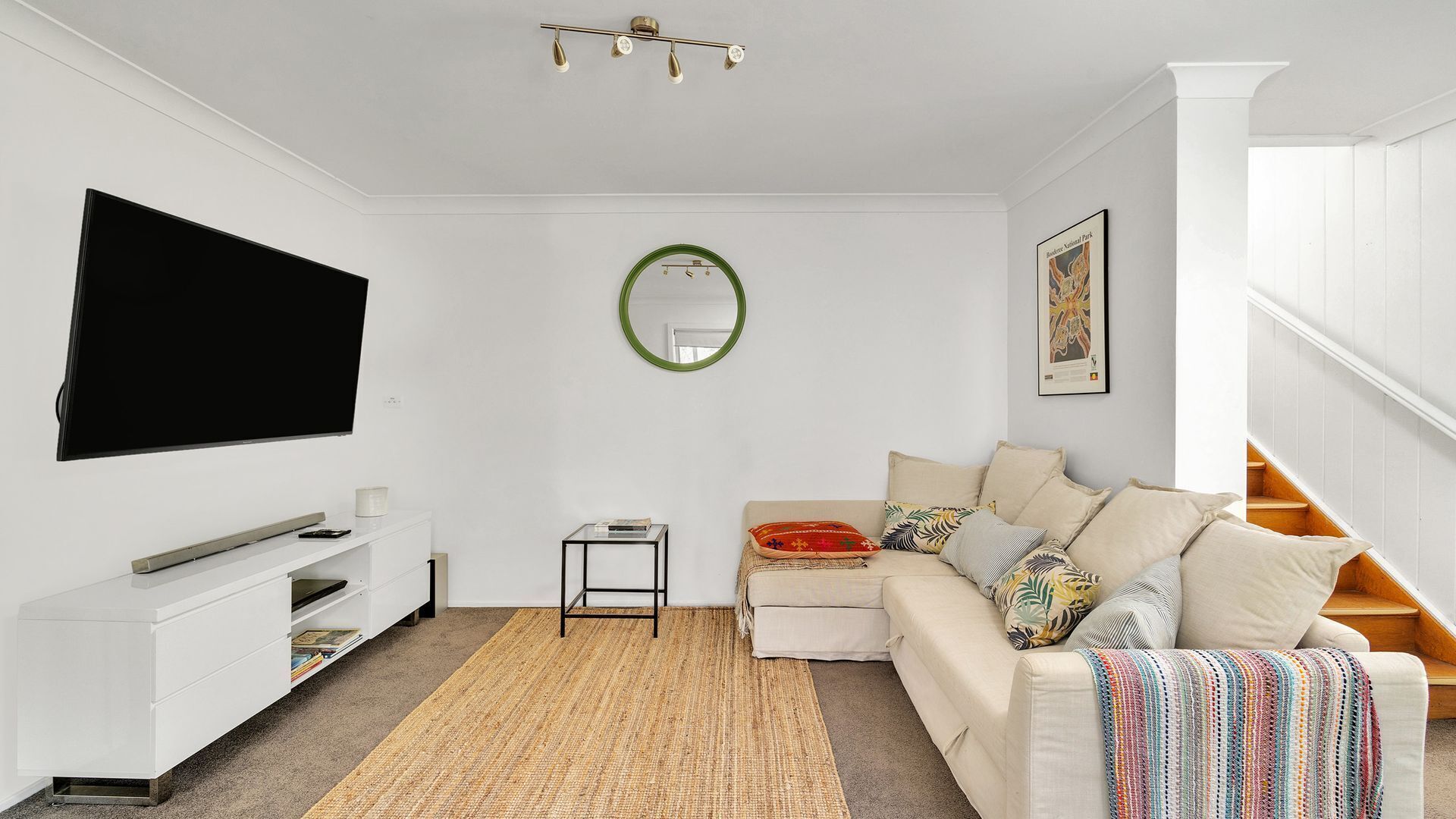 A modern living room with a flat-screen TV, a white sofa, a small side table, a woven rug, and a staircase with wooden steps.