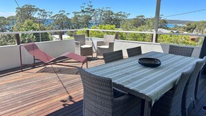 A deck with wicker furniture, a table with a bowl, and a view of the sea and trees. - Unwind at Hyams Beach, Jervis Bay (Hyams Beach)