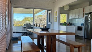 A modern kitchen with white cabinets, a stainless steel refrigerator, and a dining area with a wooden table and bench. - Unwind at Hyams Beach, Jervis Bay (Hyams Beach)