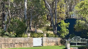 A stone wall with a metal gate, a bench, and a small garden area with various plants and trees. - Unwind at Hyams Beach, Jervis Bay (Hyams Beach)