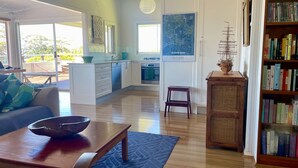A modern living room with a large window, a dining area with a bowl on a table, a kitchen with white cabinets, and a bookshelf filled with books. - Unwind at Hyams Beach, Jervis Bay (Hyams Beach)