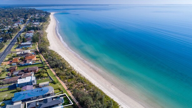 Oversigtsbillede af et kystnære boligområde med huse langs stranden, et klart blåt hav og en vej parallelt med stranden.