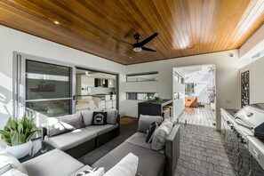 A modern living room with a wooden ceiling, a ceiling fan, and a large sliding glass door leading to a kitchen area. - ULTRA STYLISH FREMANTLE AREA WALK TO THE BEACH (Beaconsfield)