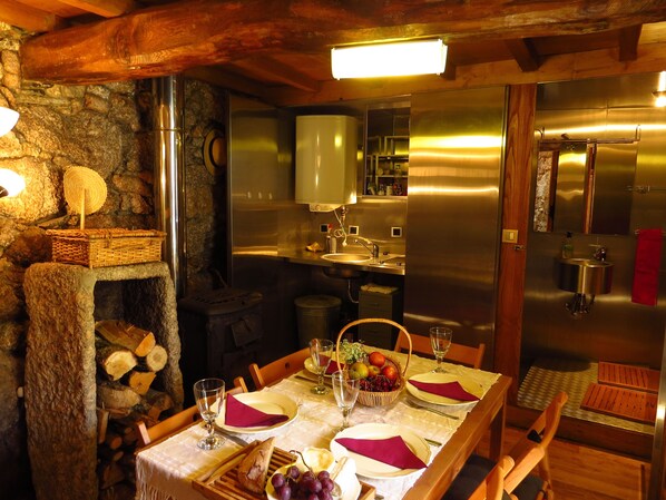 A rustic dining area with a stone wall, wooden beams, a chandelier, a buffet table, and a modern kitchen in the background. - Room in Cabin - Restored, Rustic and Rural Mini Cottage in Typical Portuguese Village (Lamego)