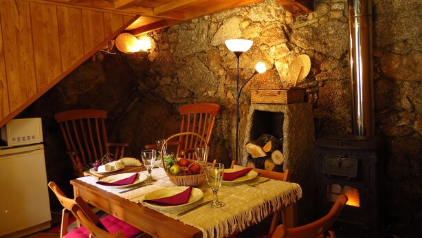 A cozy dining area with a stone wall, wooden ceiling beams, and a wood-burning stove. - Room in Cabin - Restored, Rustic and Rural Mini Cottage in Typical Portuguese Village (Lamego)