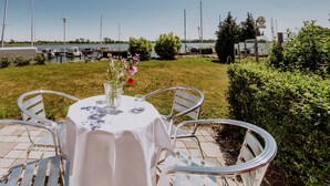 Un área de comedor al aire libre con una mesa preparada para dos personas, rodeada de sillas de metal, con vista a un puerto deportivo lleno de barcos.