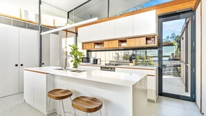 Uma cozinha moderna com ilha central, banquetas, micro-ondas, cafeteira e vista para o exterior através de uma porta de vidro.
