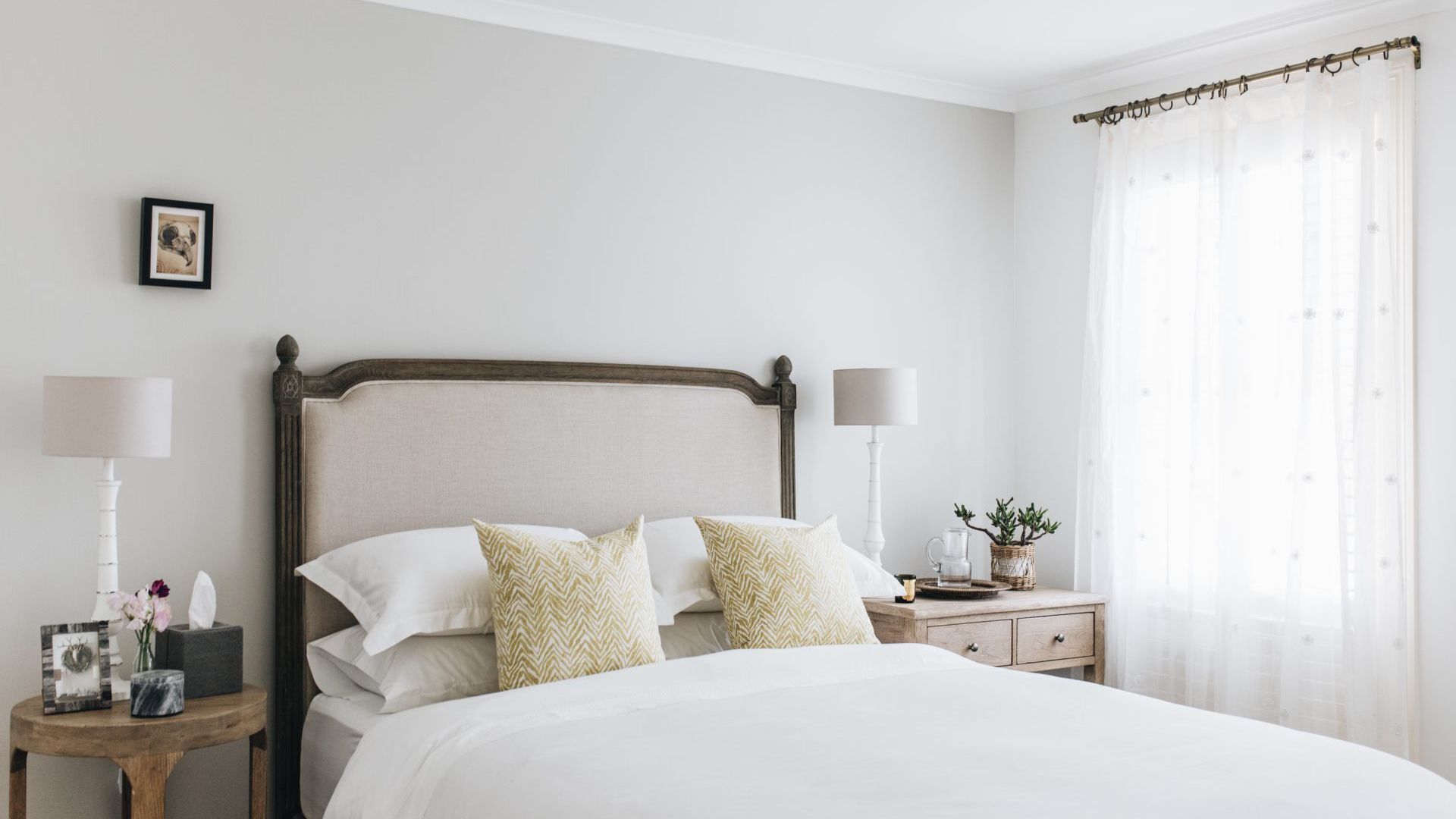 A neatly arranged bedroom with a bed, two bedside tables, a nightstand, a lamp, a picture frame, and a window with sheer curtains.