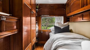 A narrow, wooden-paneled train compartment with a bed, pillows, and a window with a view of greenery. A 'NON-SMOKING' sign is visible above the window.