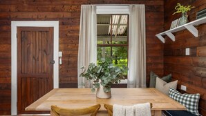 A wooden interior with a dining table, chairs, a vase with greenery, and a window with curtains.