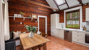A rustic kitchen with wooden walls, a wooden dining table, and a window overlooking a garden.