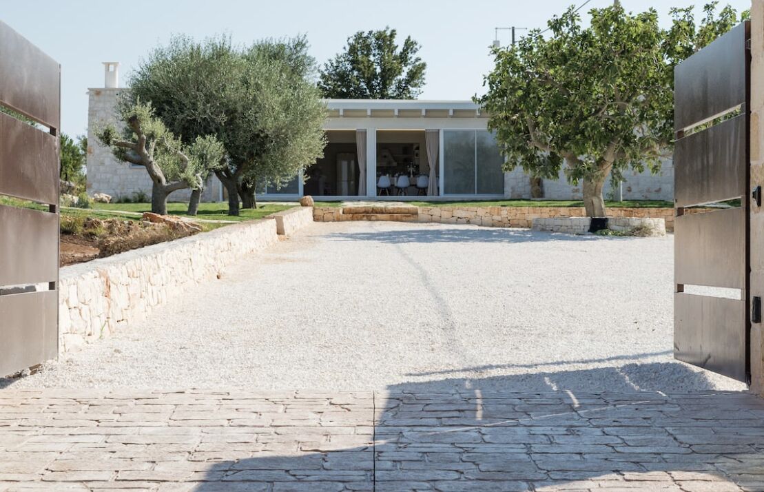 Ein gepflasterter Weg führt zu einem modernen Haus mit Steinmauer und Olivenbäumen.