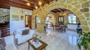 A spacious living area with a white sofa, a coffee table with a vase and flowers, a stone archway, a kitchen in the background, and a wooden ceiling. - Traditional Fully Private Villa with Pool (Chania)