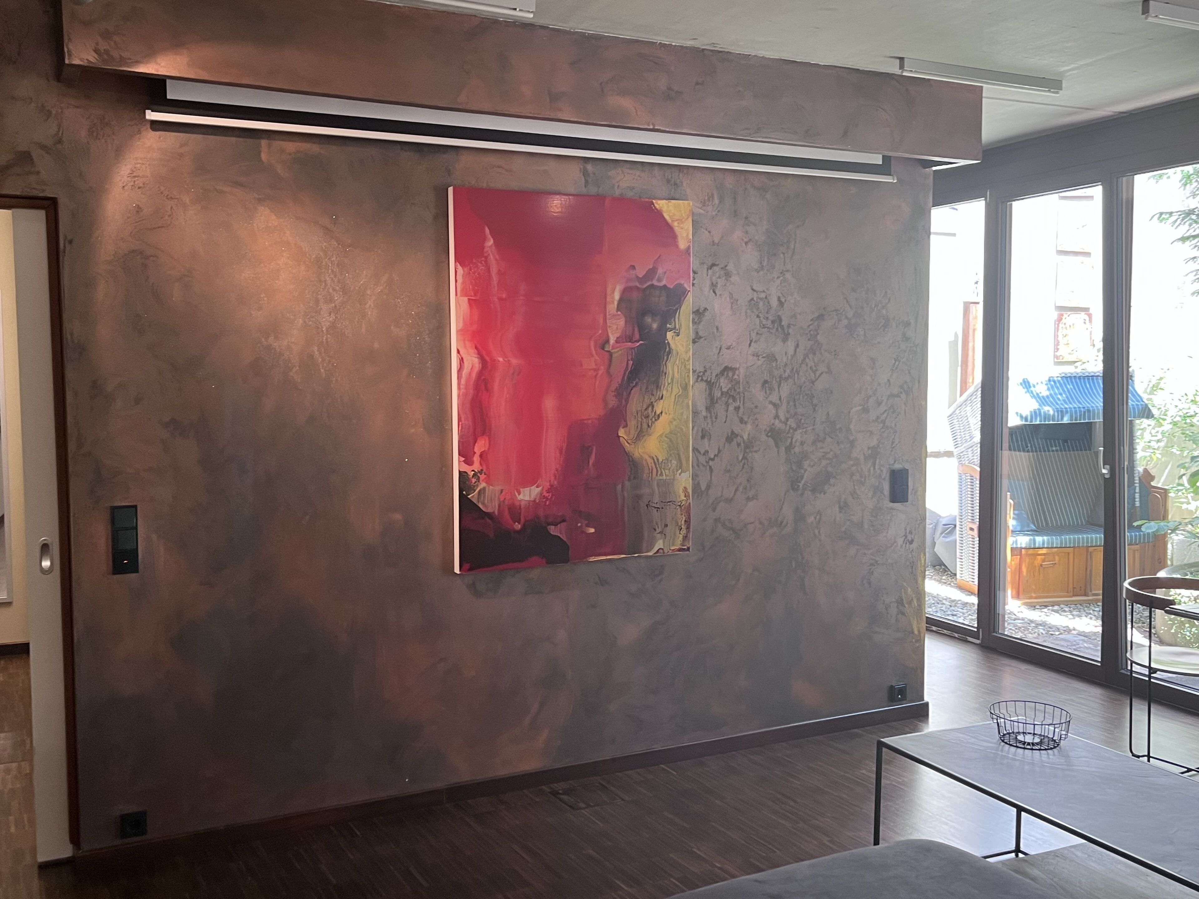 A modern interior with a large abstract painting on a textured wall, a sliding glass door leading to an outdoor area, and a minimalist table with a wire basket.