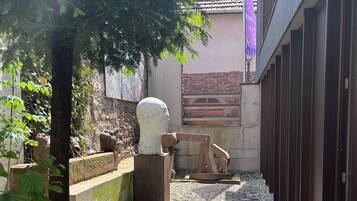 A narrow pathway leading to a building with a tiled roof and a stone wall. There is a tree on the left and a purple flag on the right.