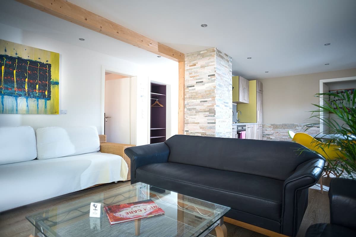 Una sala de estar moderna con un sofá blanco, un sofá de cuero negro y una mesa de centro de vidrio. En la pared, un cuadro abstracto de colores vivos, y se ve un área de cocina al fondo.