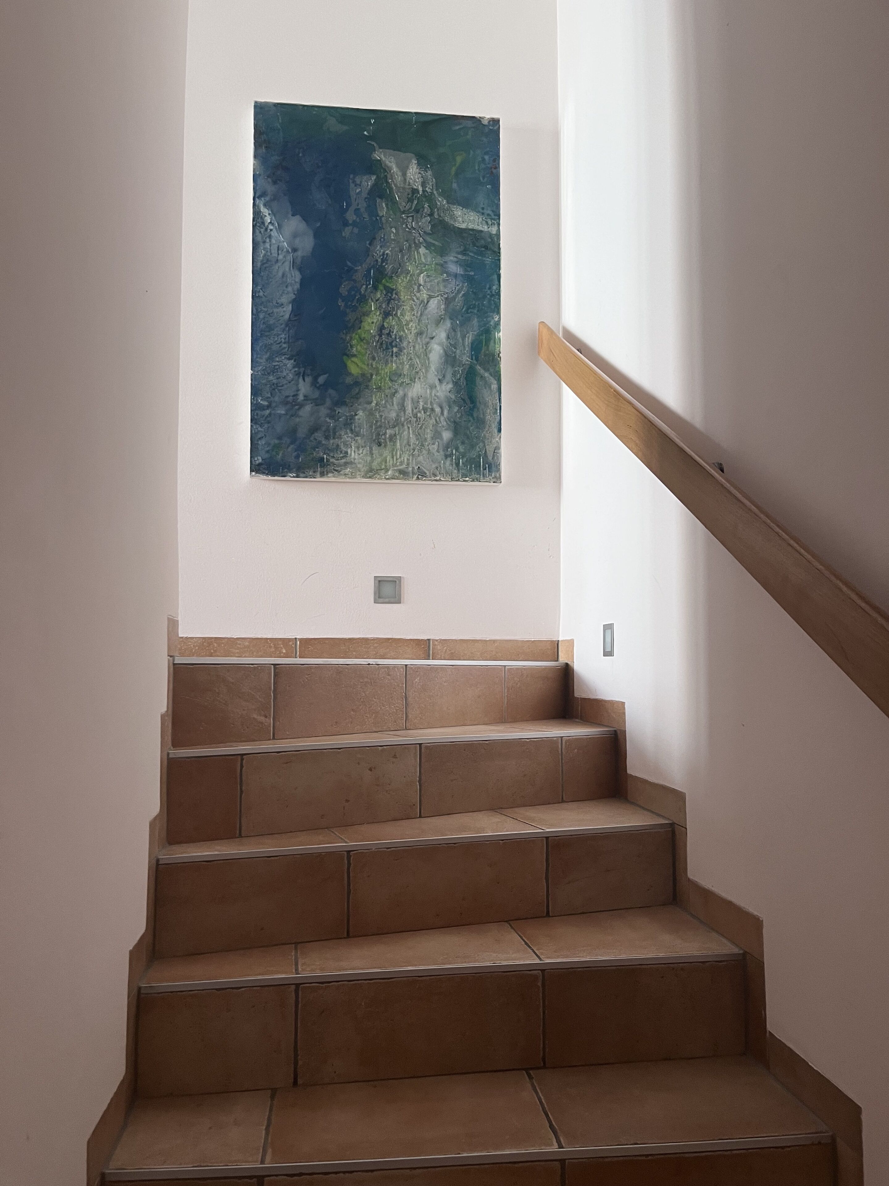 Un escalier recouvert de carreaux bruns et doté d’une rampe en bois, menant à un mur orné d’un grand tableau abstrait.