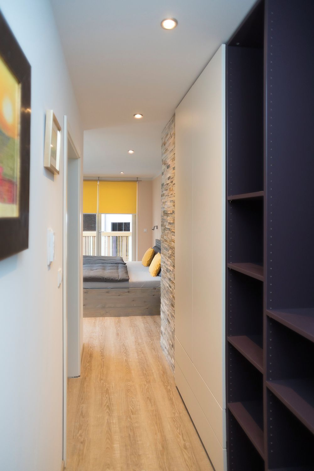 A modern hallway with a wooden floor, a stone accent wall, and a bed with yellow pillows.