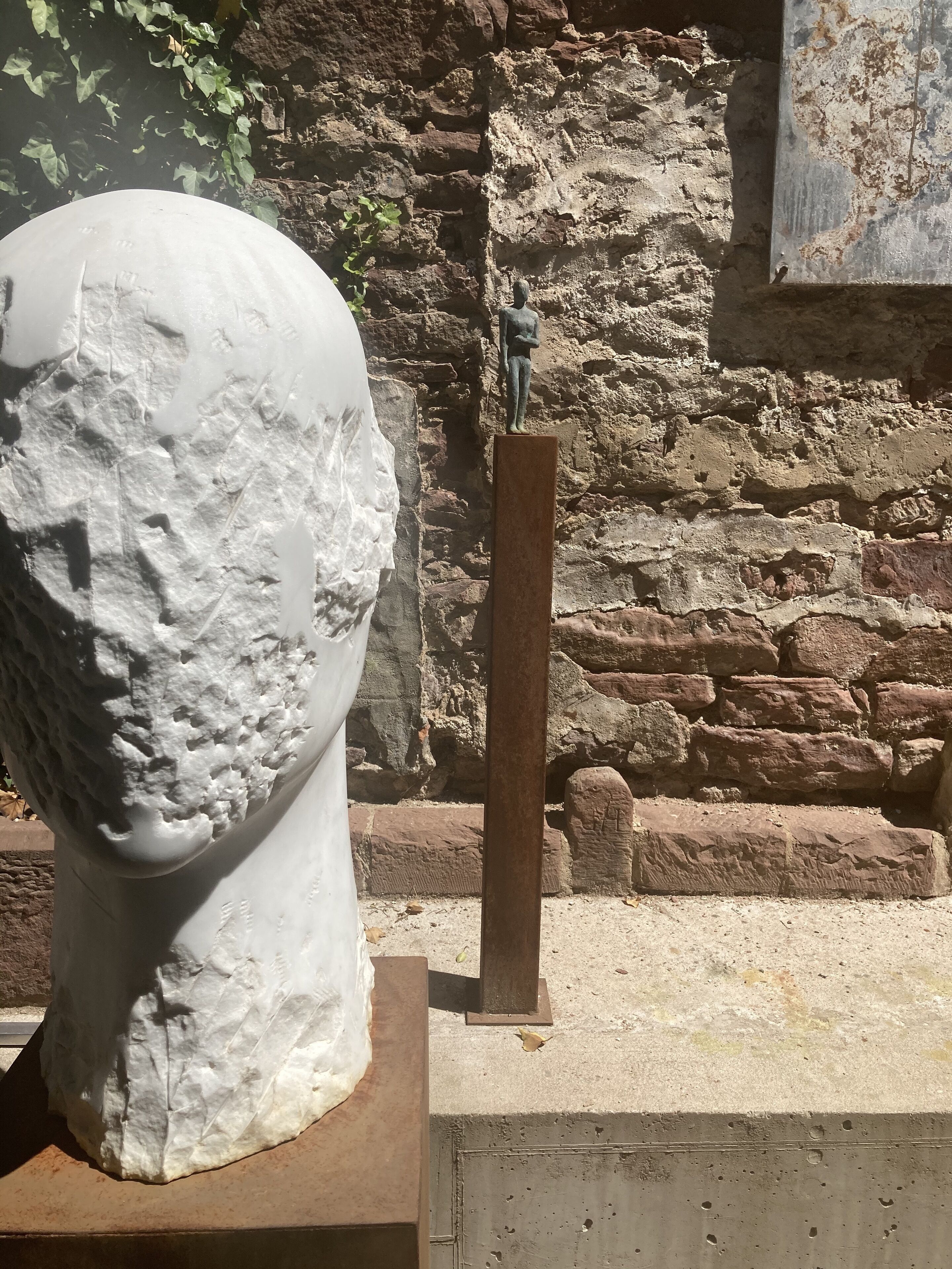 A white bust sculpture on a pedestal, a bronze statue, and a stone wall with ivy in the background.
