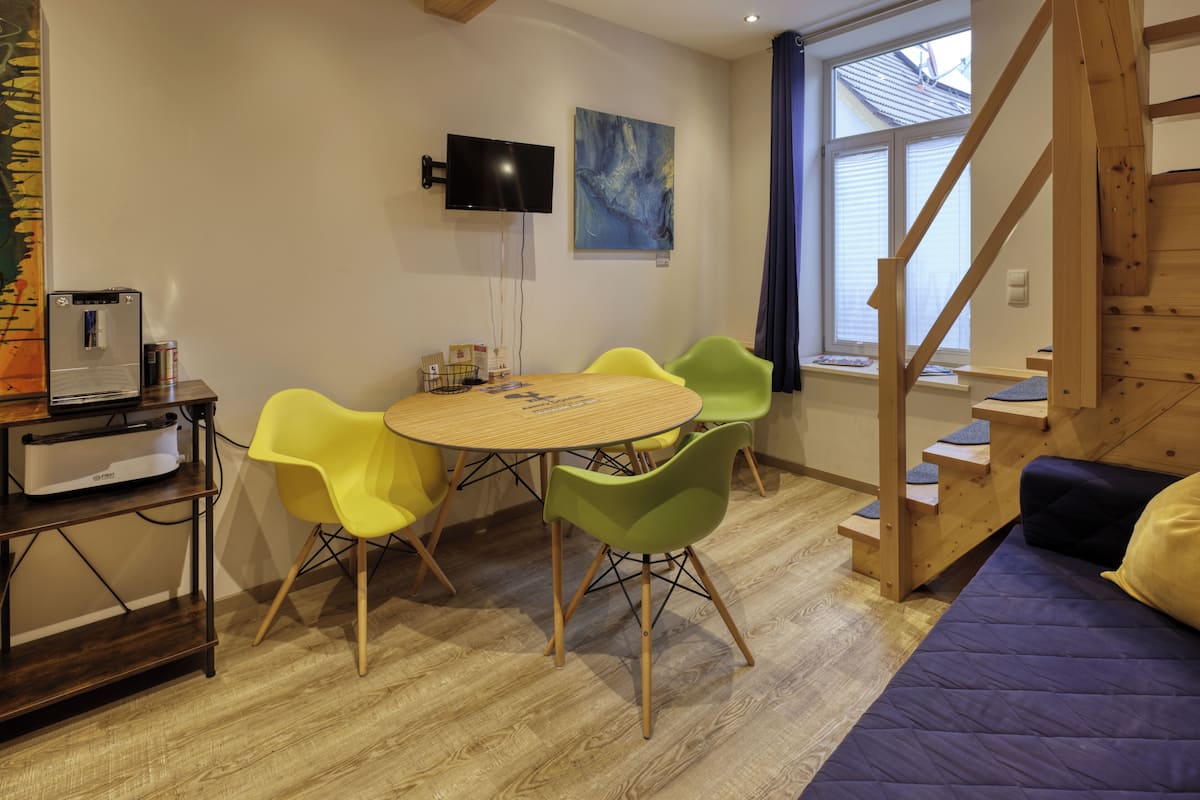 Una habitación con escalera de madera, una mesa redonda con sillas, una cama, un televisor y un ventanal con cortinas.