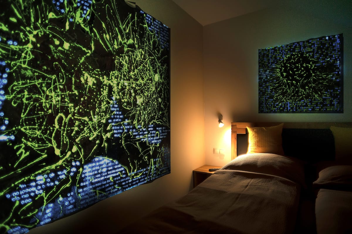 Un dormitorio con una cama, una mesita de noche y dos pantallas fijadas a la pared mostrando arte digital abstracto.