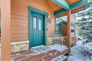 A wooden cabin with a green door, stone foundation, and a porch with a chair.