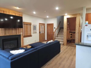 A living room with a fireplace, a large flat-screen TV, a wooden ceiling, and a staircase leading to another level.